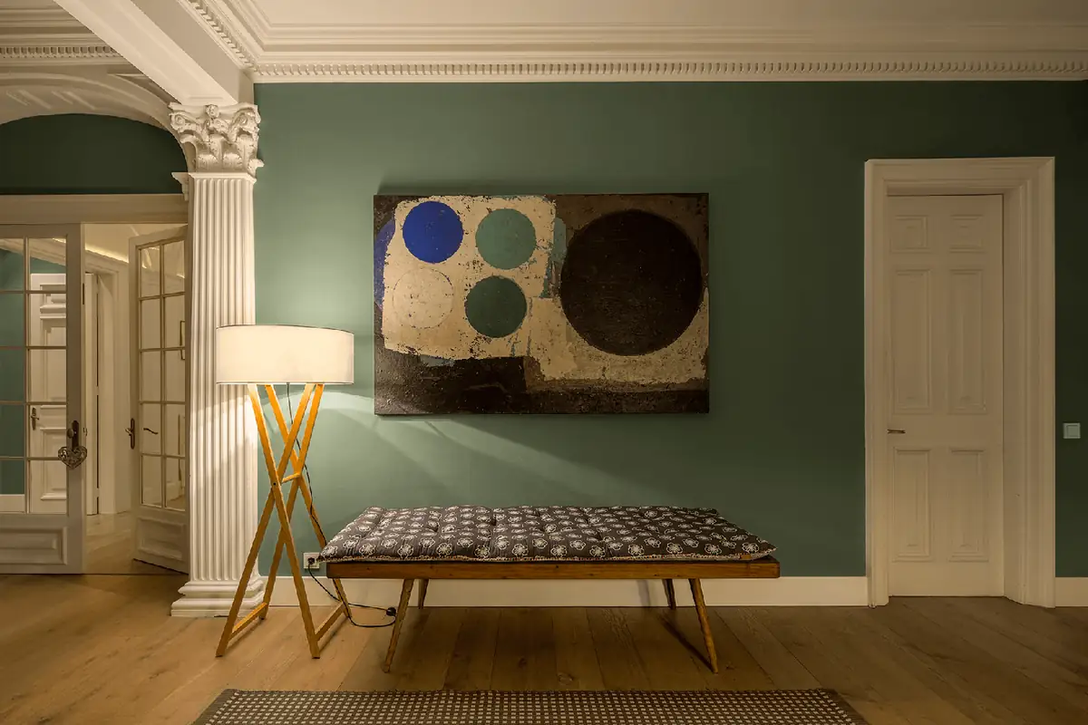 Banco de madera tapizado, lámpara de pie con trípode de madera y cuadro abstracto sobre una pared verde