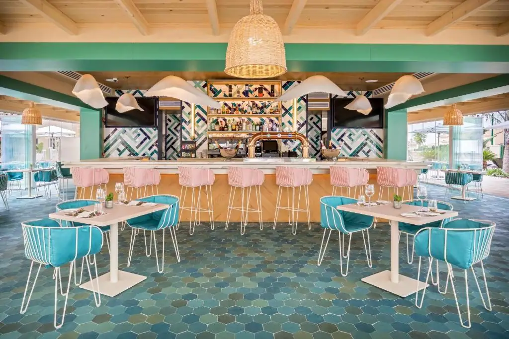 Gastrobar con barra de madera, taburetes rosas, sillas turquesa y frente de azulejos en azules, verdes y blancos