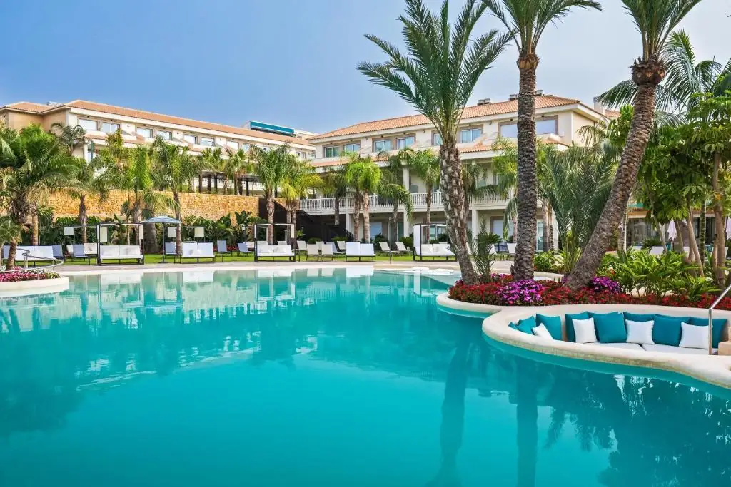 Piscina tipo lago entre palmeras con una península chill-out de sofás blancos y turquesa frente a los edificios del hotel