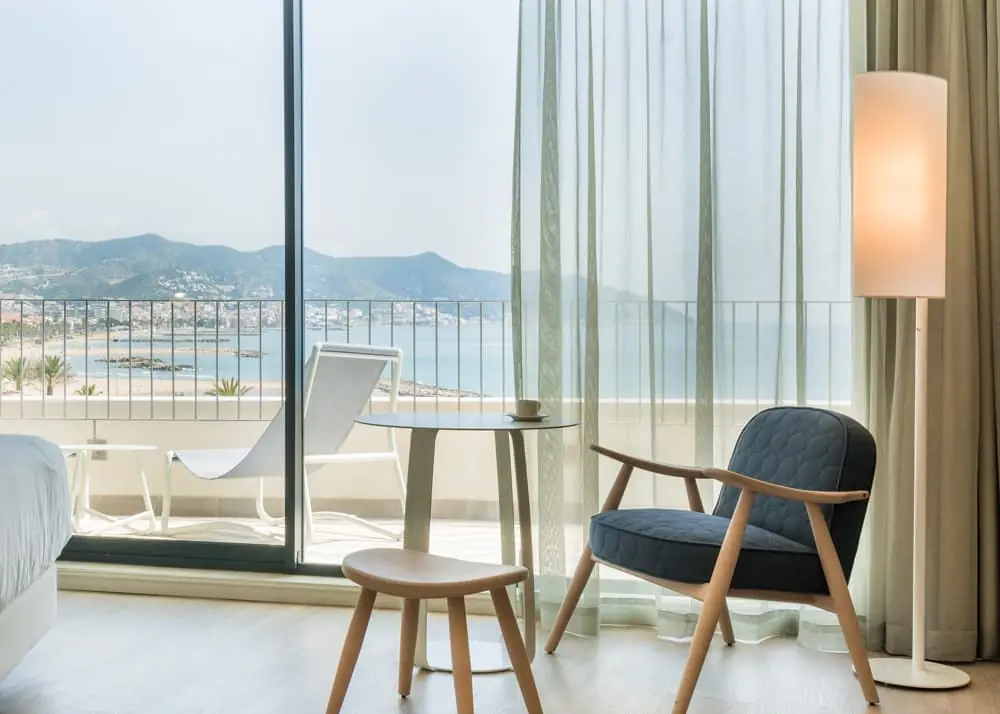 Habitación con butaca tapizada, mesa redonda y ventanal abierto a una terraza con vistas al mar