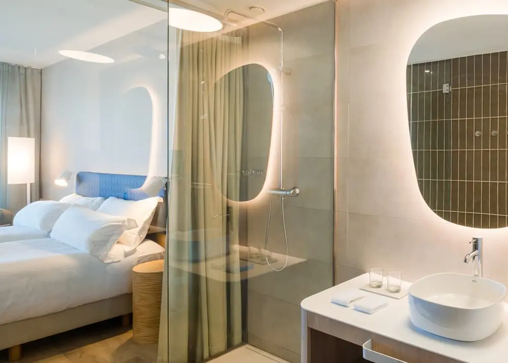 Habitación con baño integrado tras una mampara de vidrio, lavabo sobre encimera y espejos retroiluminados