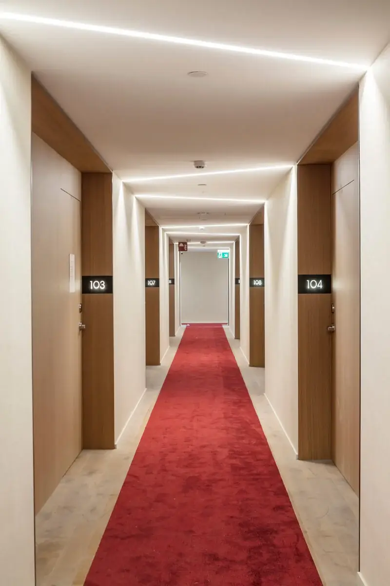Pasillo de habitaciones con alfombra roja, marcos de puertas de madera y línea de luz en el techo