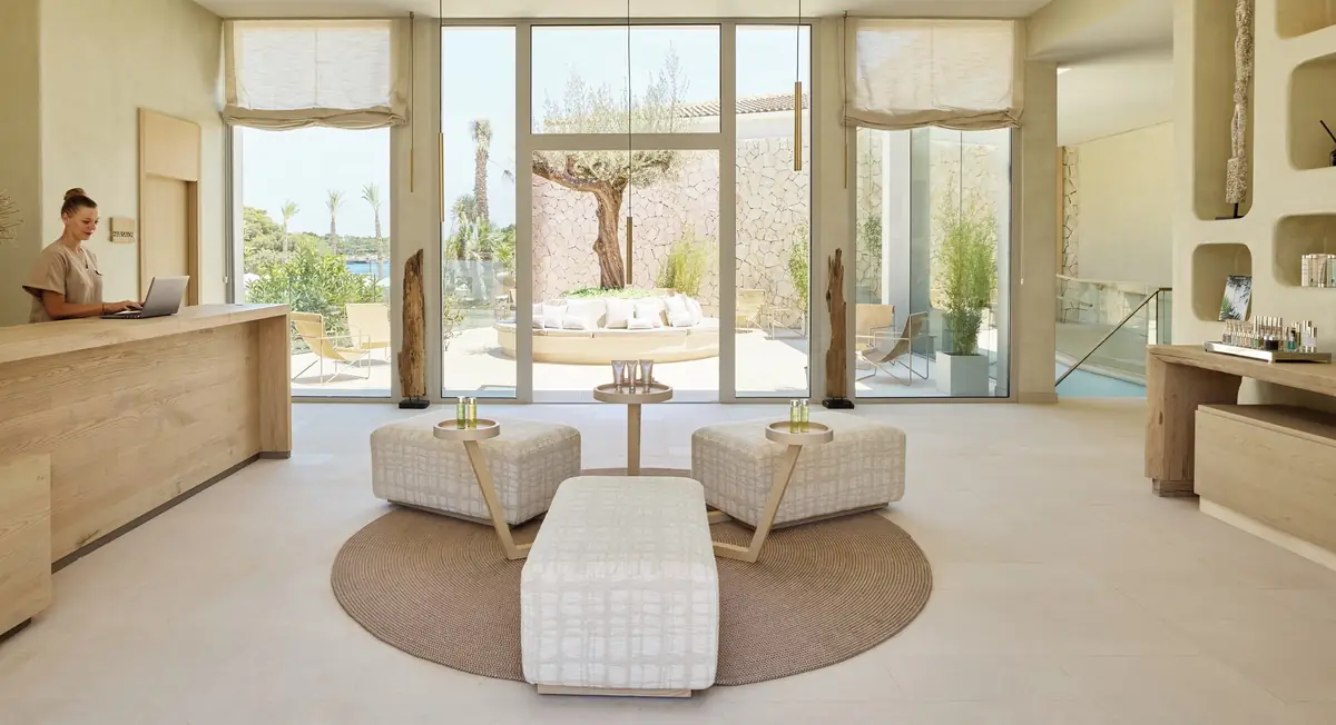 Recepción del spa con mostrador de madera clara, pufs blancos y ventanales al jardín