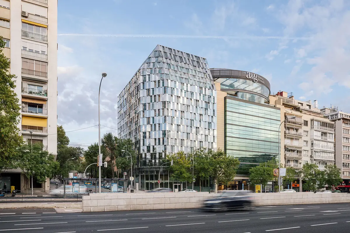 Vista del edificio Castellana 94 desde el paseo, con su volumen facetado de vidrio entre edificios vecinos y arbolado