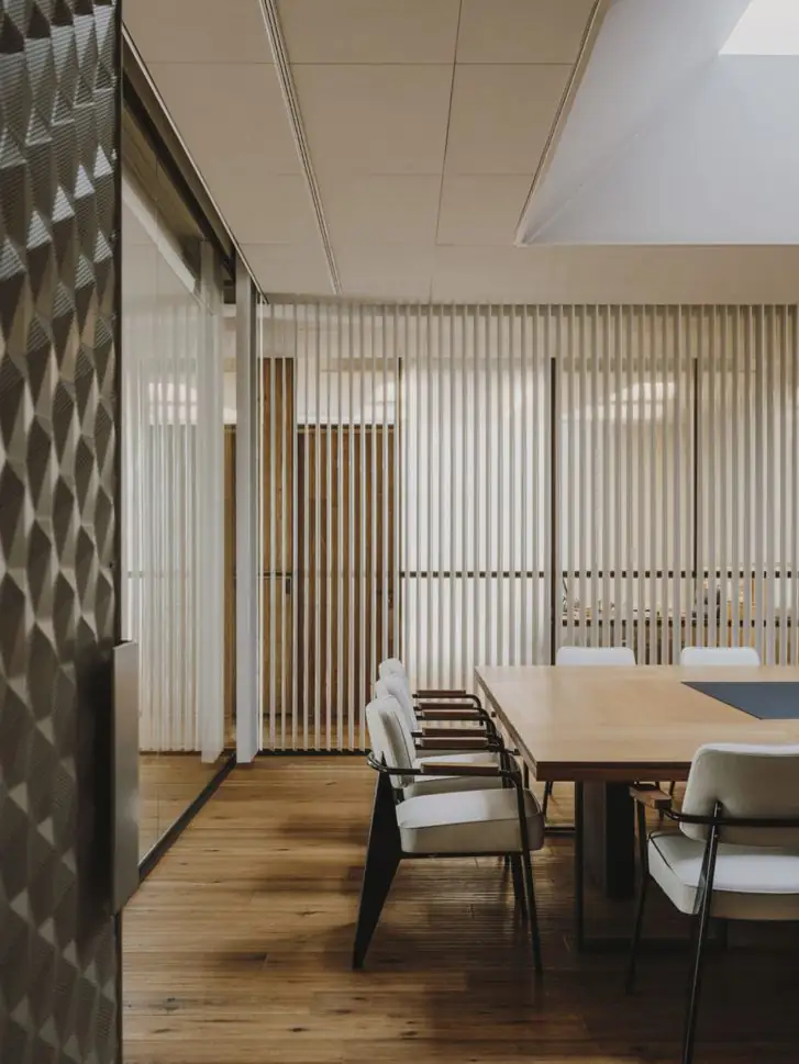 Mesa de reuniones con sillas tapizadas en blanco, cortinas de lamas y un revestimiento en relieve