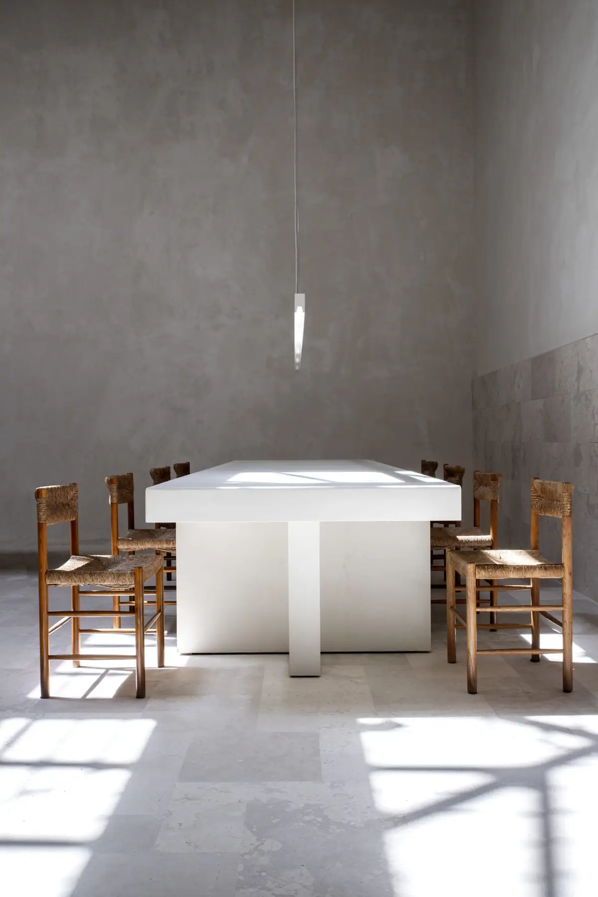 Mesa de reuniones blanca con sillas de madera y asiento de enea bajo una lámpara colgante, con la luz del sol en el suelo de piedra