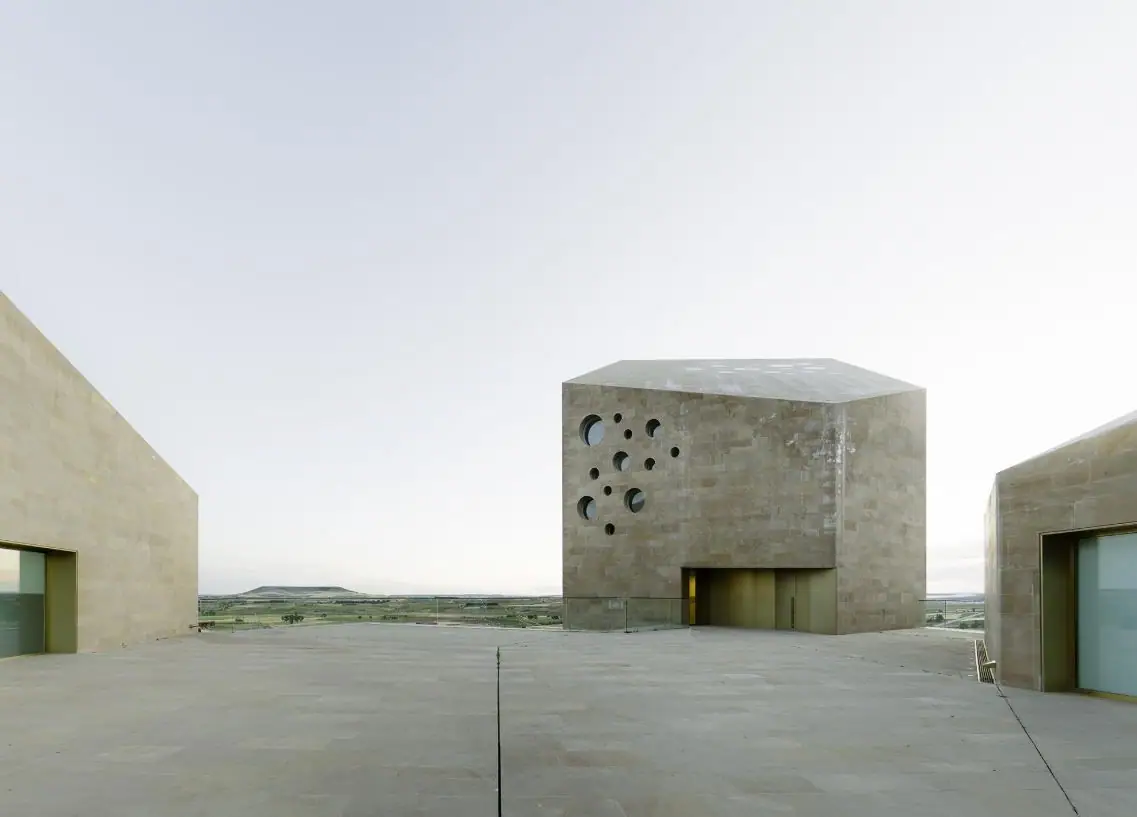 Plaza mirador de piedra entre volúmenes de sillería, con la torre de huecos circulares y la meseta al fondo