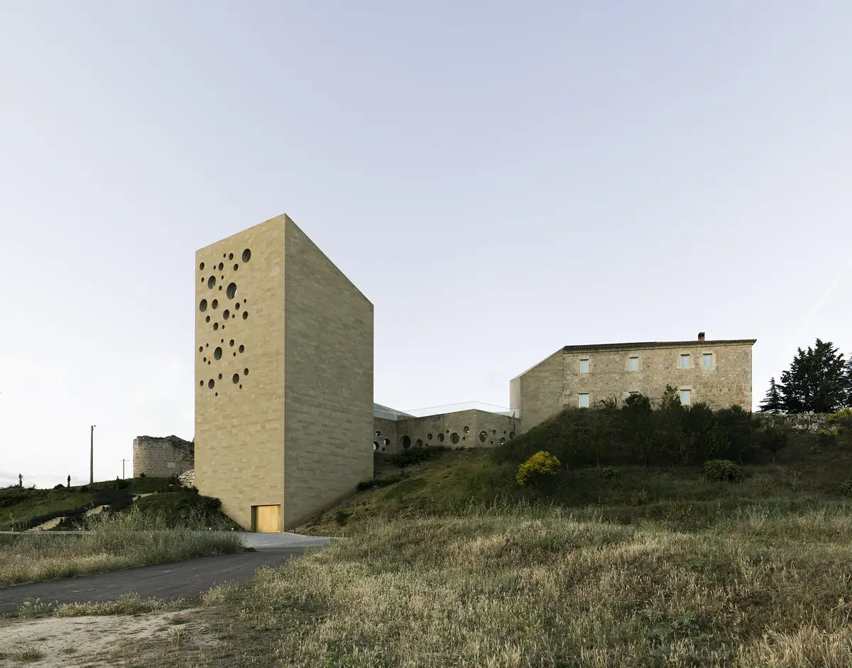 Torre de piedra ocre con aberturas circulares junto al antiguo hospital, sobre la ladera