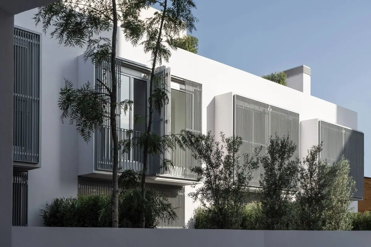Fachada interior blanca con celosías metálicas grises y árboles delante