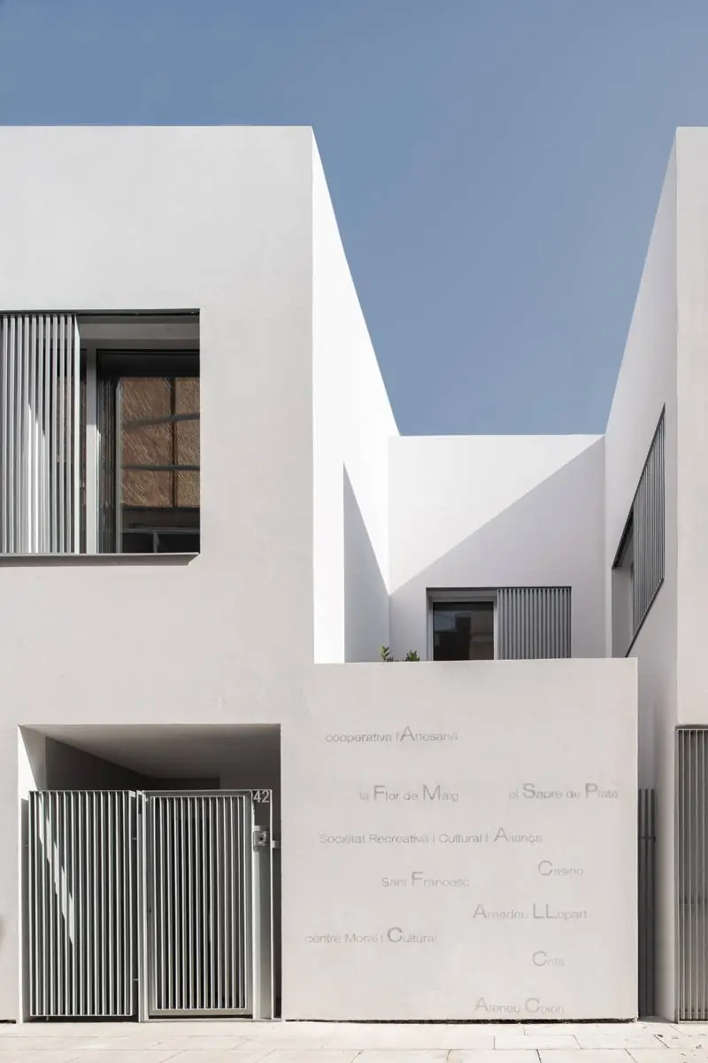 Muro blanco de planta baja con nombres del barrio pintados en gris junto a una puerta de lamas