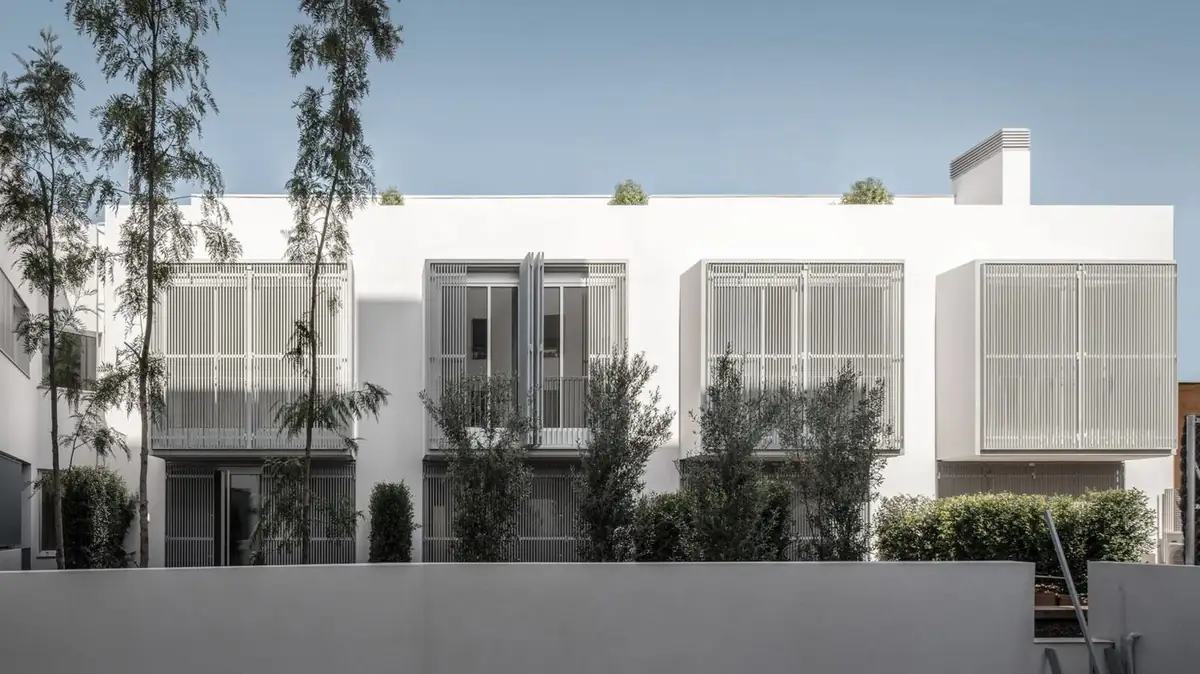 Fachada blanca de las viviendas con cuatro cuerpos de celosías y árboles en primer plano