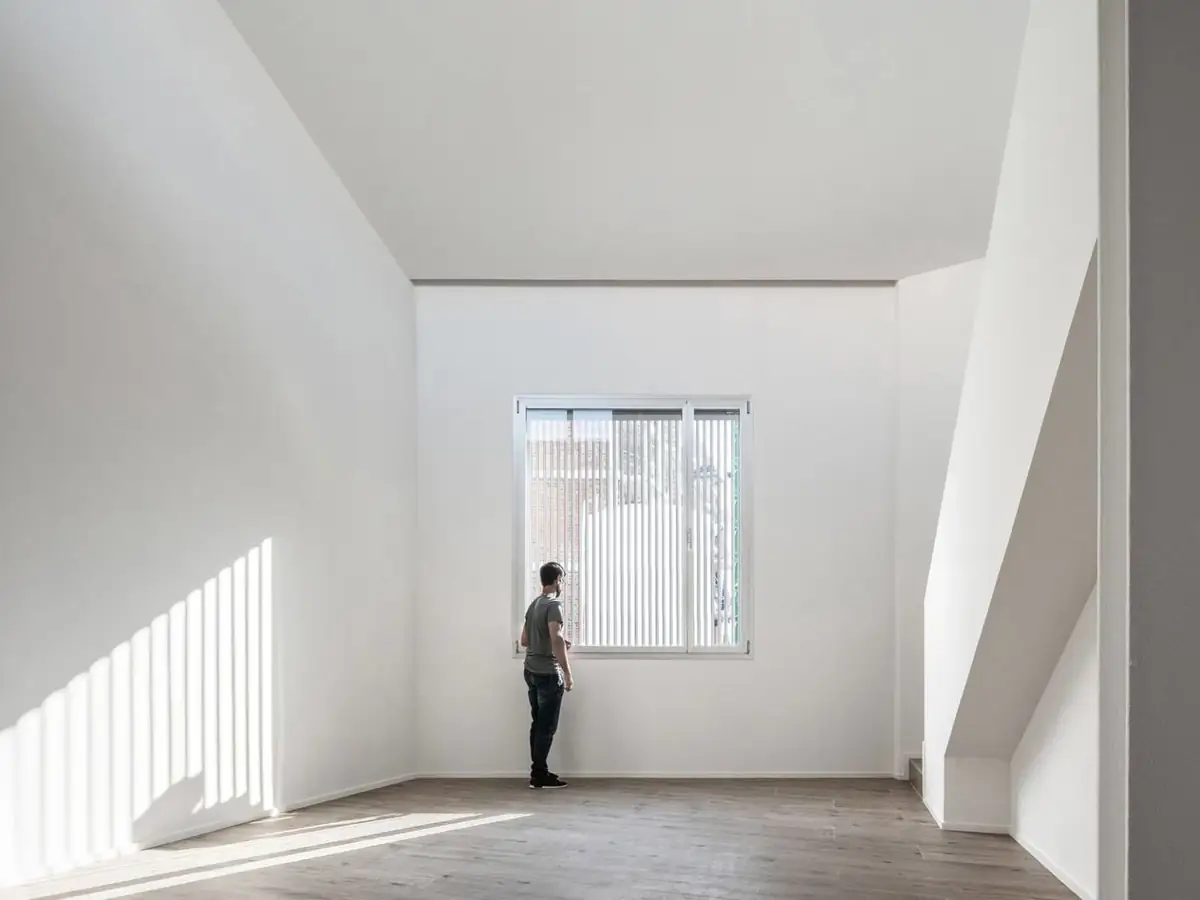 Dormitorio de doble altura con paredes blancas, ventana con lamas y luz rayada en la pared