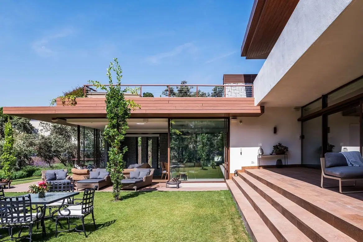 Porche de una vivienda unifamiliar con cubierta de madera volada, cerramientos de vidrio, escalinata cerámica y jardín