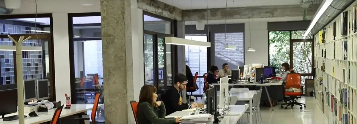 Puestos de trabajo en una planta diáfana con pilares de hormigón, lámparas colgantes y una larga estantería de libros