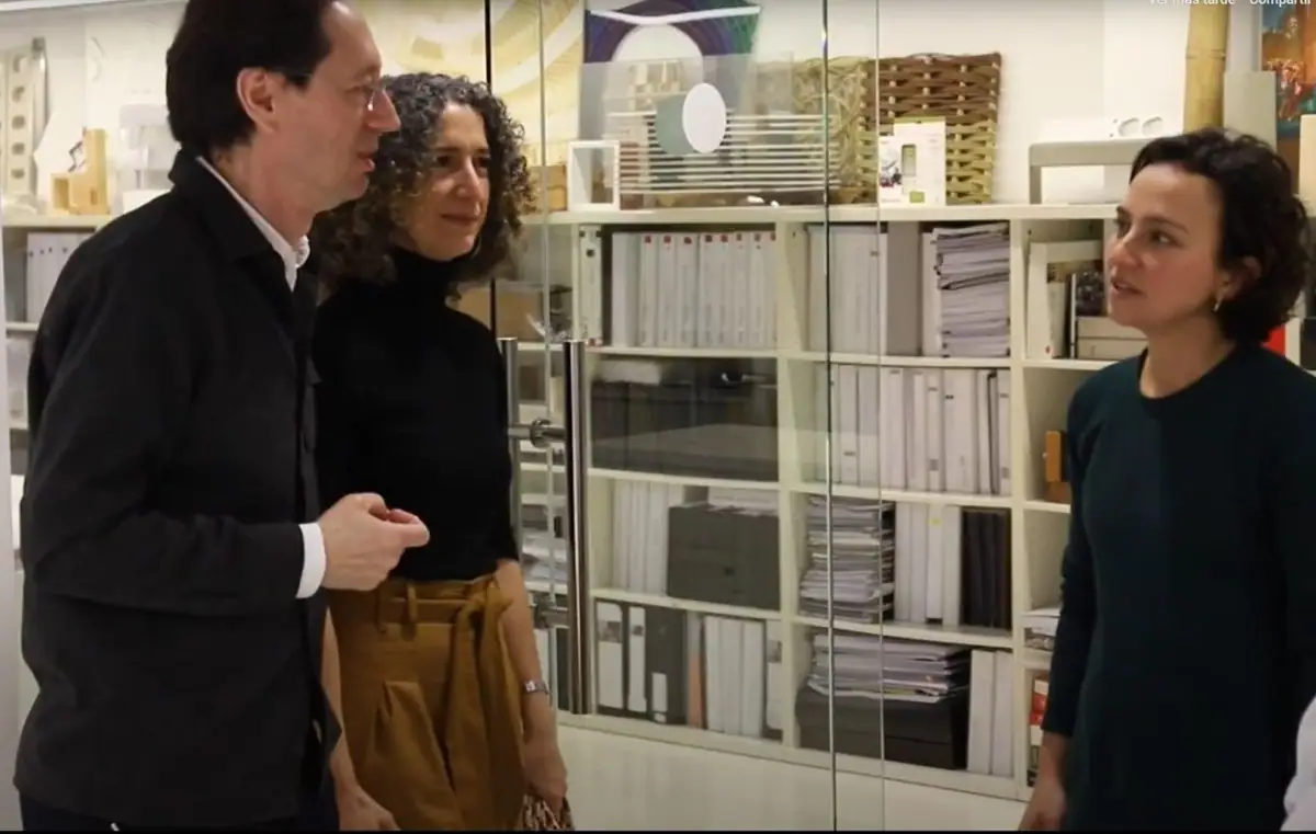 Fermín Vázquez y Ana Bassat conversan de pie con una entrevistadora frente a una estantería de maquetas y archivadores