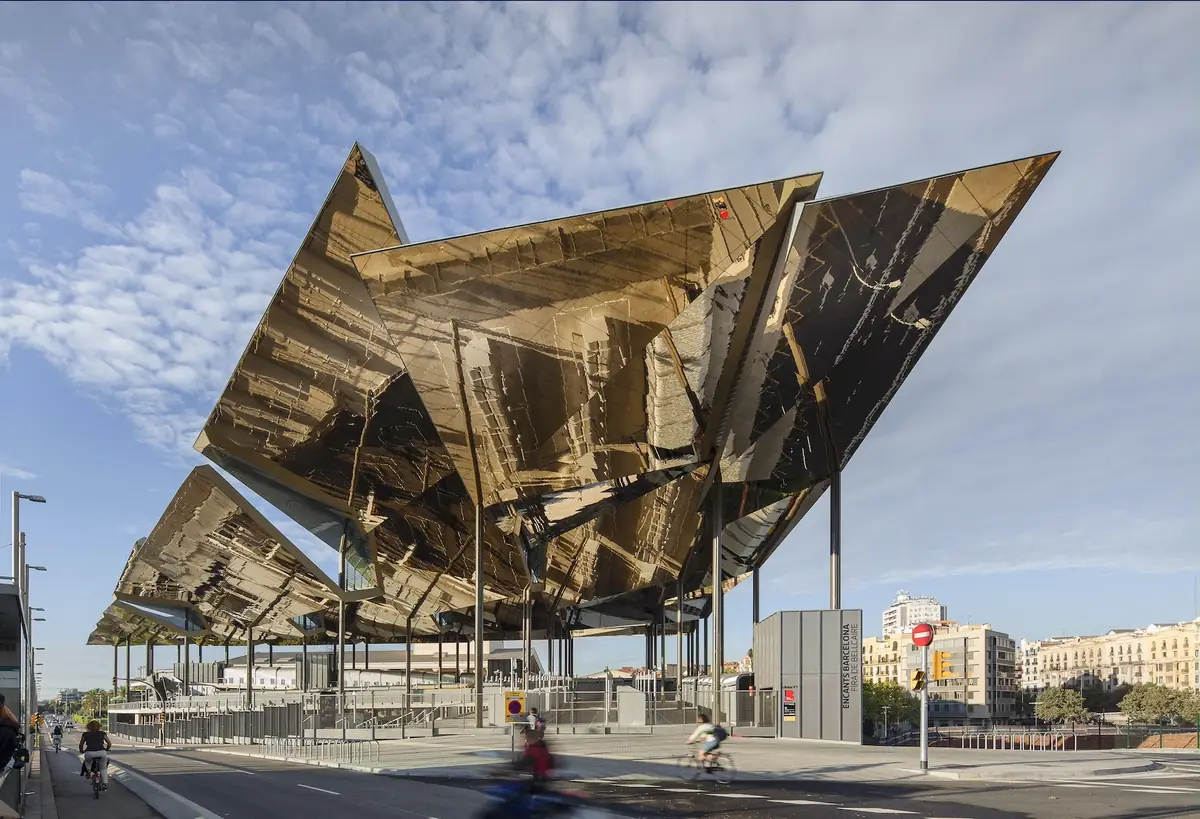 Gran cubierta de paneles metálicos dorados y reflectantes del Mercat dels Encants sobre la calle, con ciclistas y edificios de viviendas al fondo