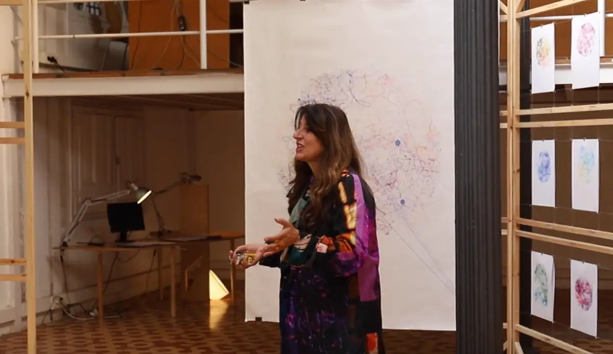 Benedetta Tagliabue habla de pie ante un gran dibujo colgado y una estructura de listones de madera con dibujos
