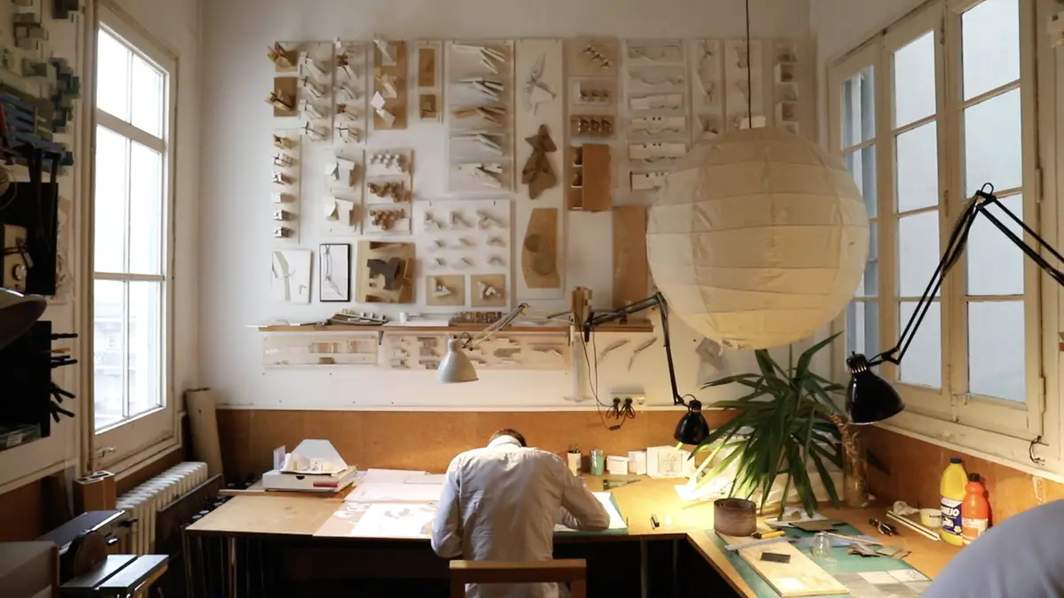 Arquitecto dibujando en una mesa bajo una pared llena de maquetas de estudio