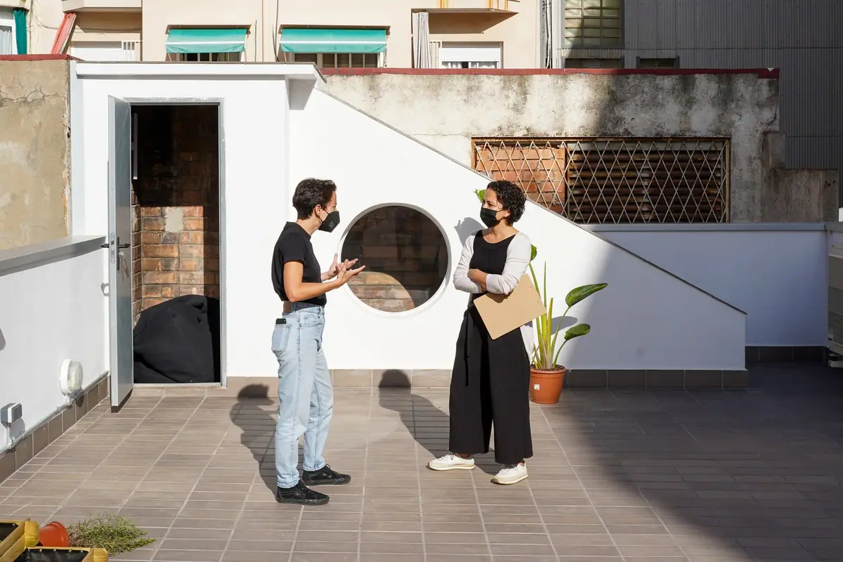 Dos mujeres con mascarilla conversan en una azotea, frente a un volumen blanco con una ventana circular