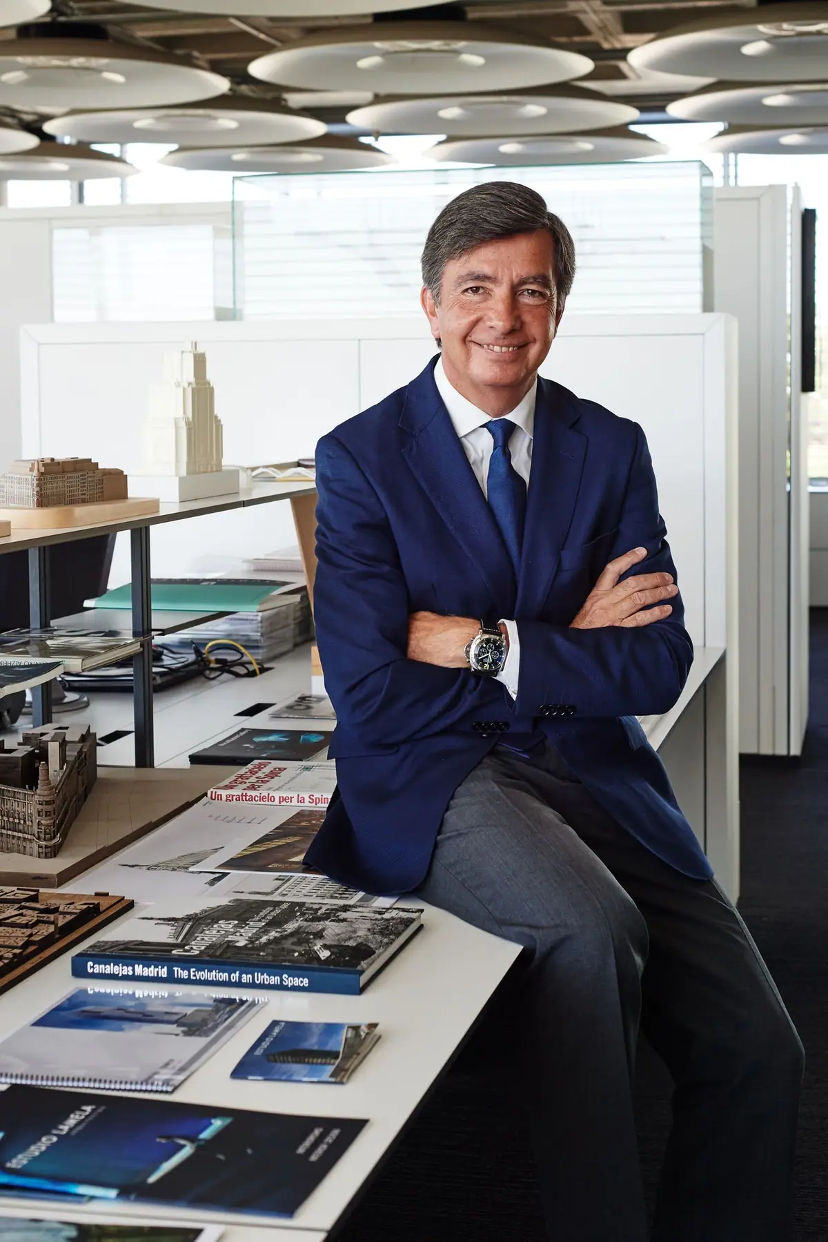 Retrato de Carlos Lamela sentado en una mesa con maquetas y publicaciones del estudio