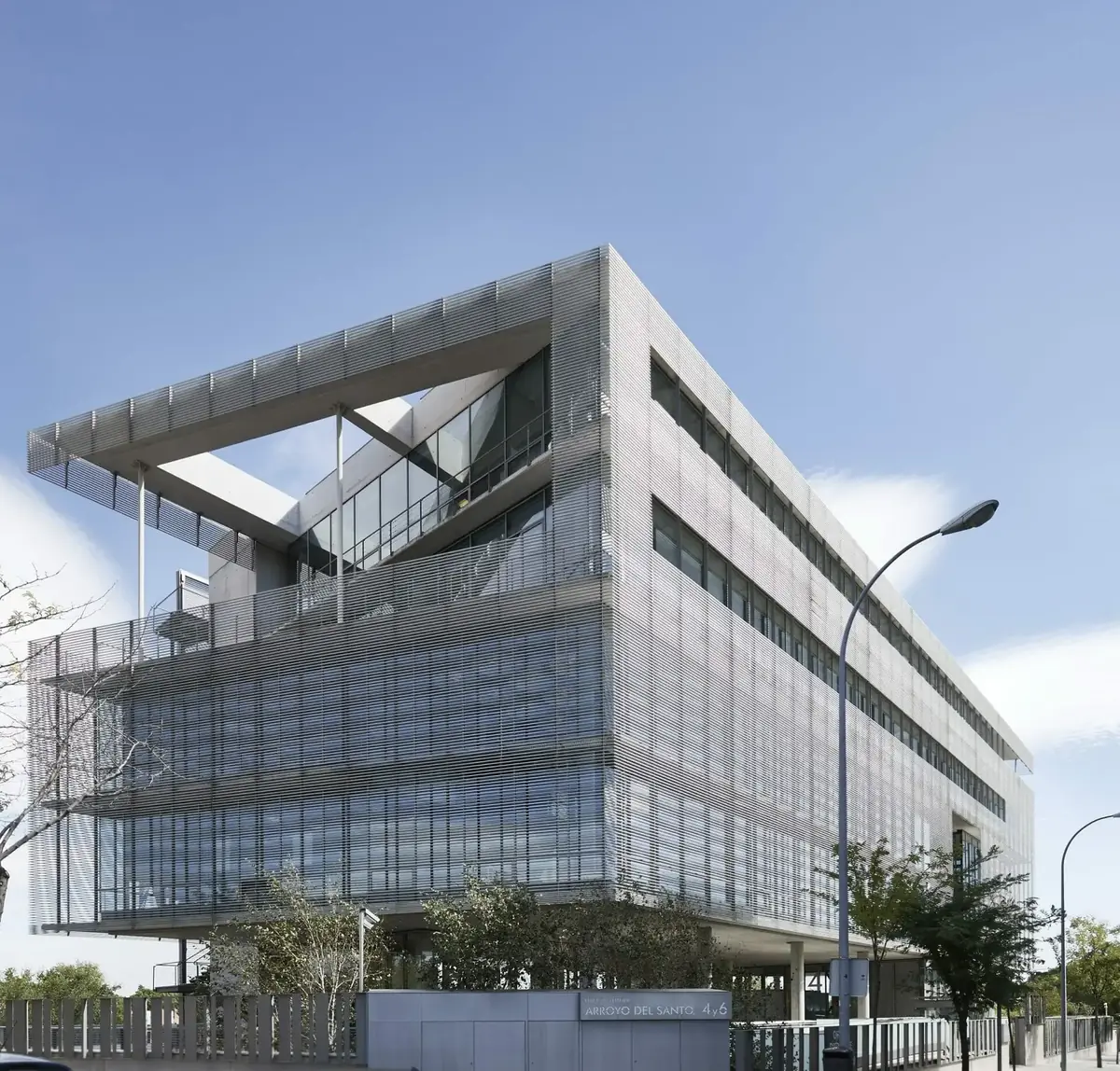 Edificio de oficinas de vidrio y malla metálica con una gran visera superior, sobre un jardín
