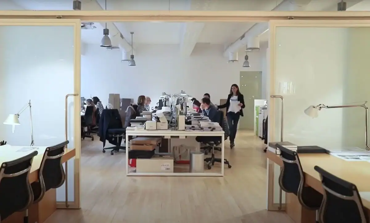 Planta de trabajo diáfana con mesas compartidas, vista a través de dos puertas de vidrio