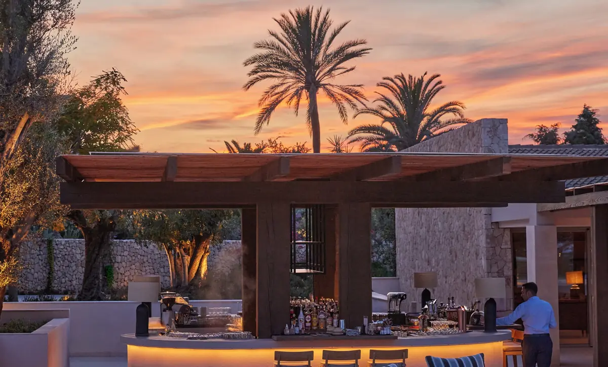 Barra exterior iluminada bajo una pérgola de madera, con muros de piedra y palmeras al atardecer