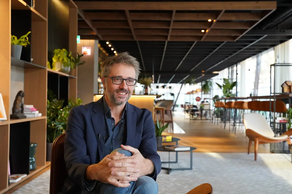 Ferran Tortosa, con gafas y americana azul, sentado en una zona de estar con techo de retícula de madera