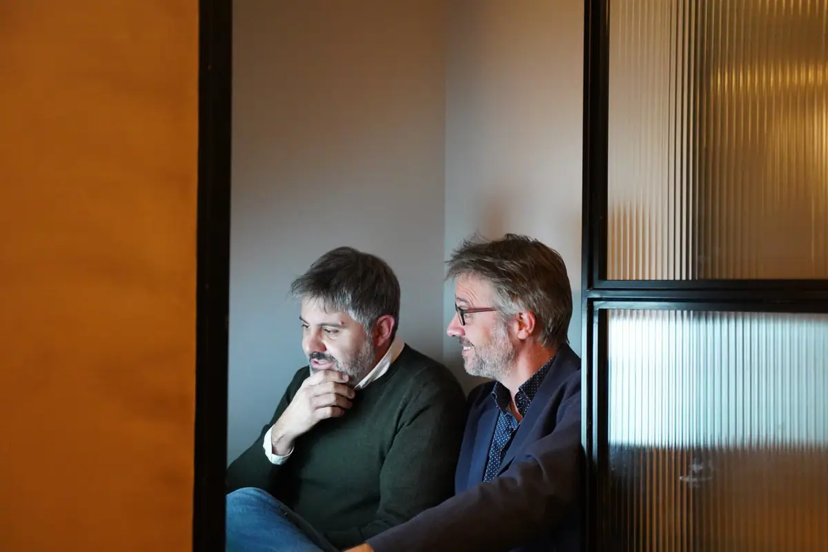 Ernest Boronat y Ferran Tortosa sentados juntos tras una puerta de vidrio acanalado, mirando hacia un lado