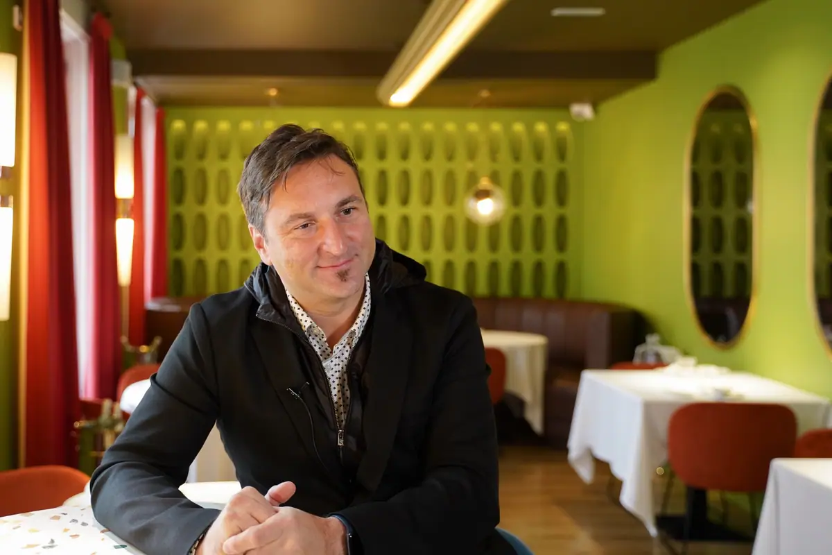 Andrea Spada sonríe sentado a una mesa en un restaurante con paredes verdes, espejos ovalados y sillas rojas