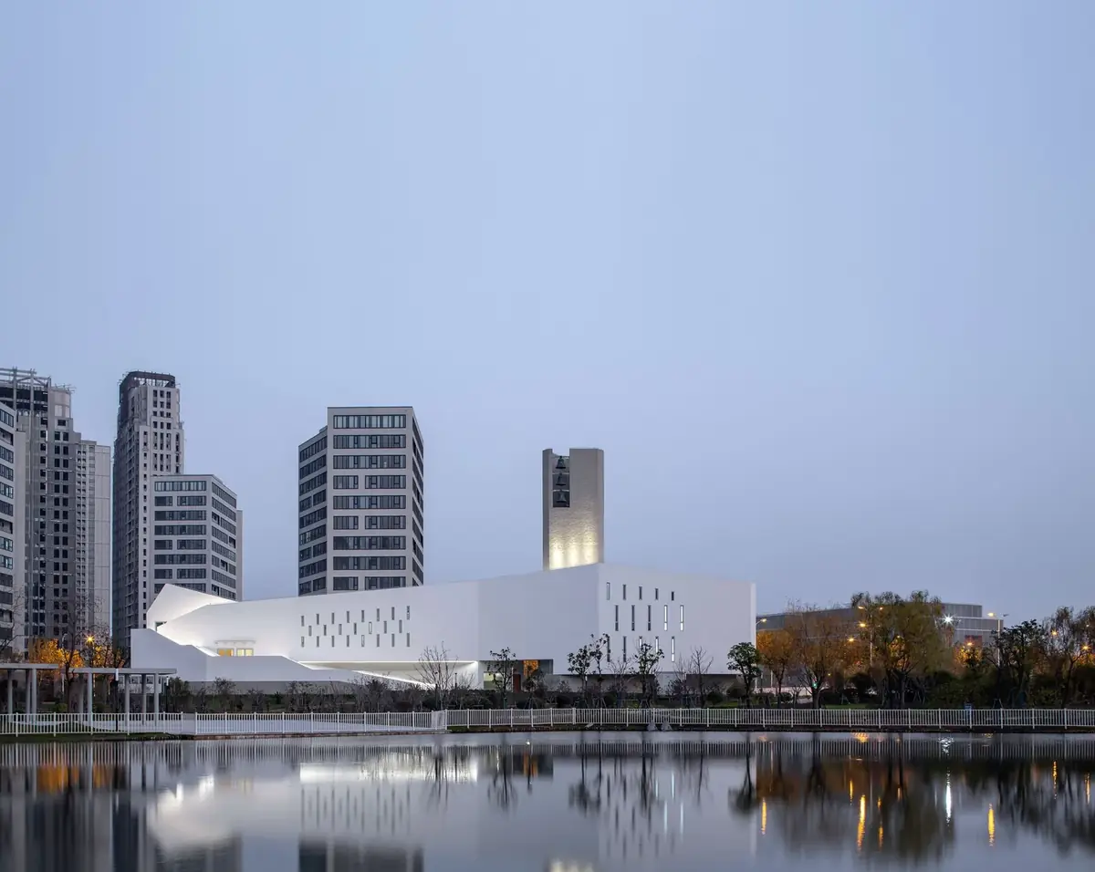 Iglesia blanca de volumen alargado y campanario reflejada en el agua al atardecer, con torres de viviendas detrás