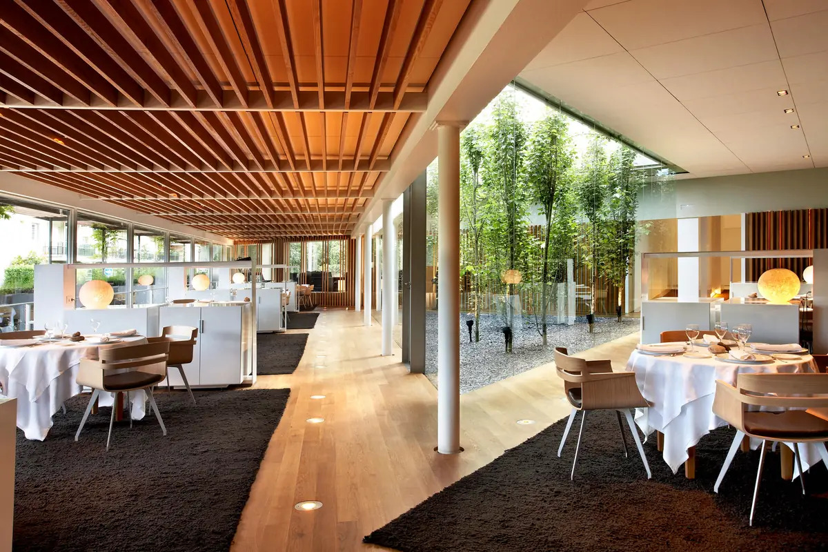 Comedor de El Celler de Can Roca con mesas vestidas de blanco, techo de listones de madera y patio acristalado con árboles
