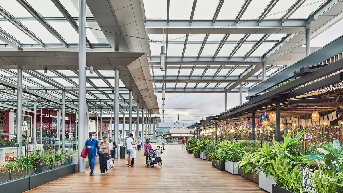 Paseo exterior de madera cubierto con pérgolas de vidrio y jardineras en el centro comercial La Maquinista, con visitantes paseando
