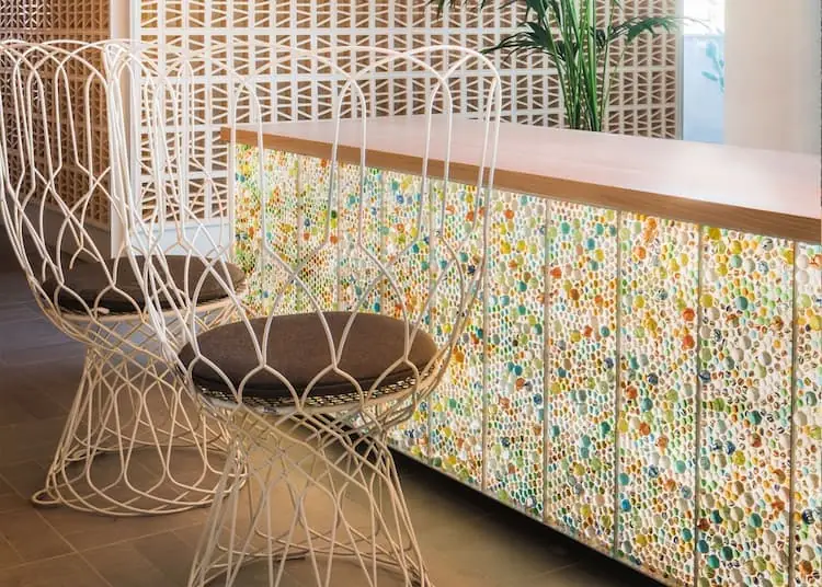 Mostrador de recepción con frente de canicas de colores retroiluminadas, encimera de madera y sillas de alambre blanco