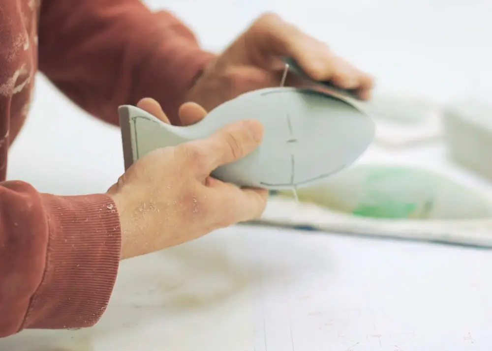 Manos con restos de polvo blanco trabajando una pieza clara en forma de pez sobre una mesa de taller