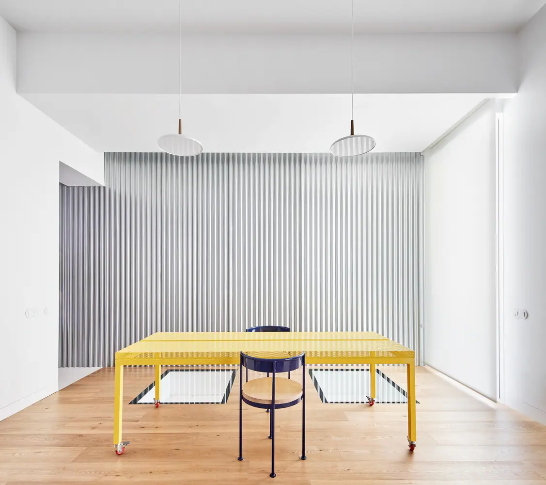 Mesa amarilla de chapa perforada con ruedas y silla azul ante una cortina metálica ondulada