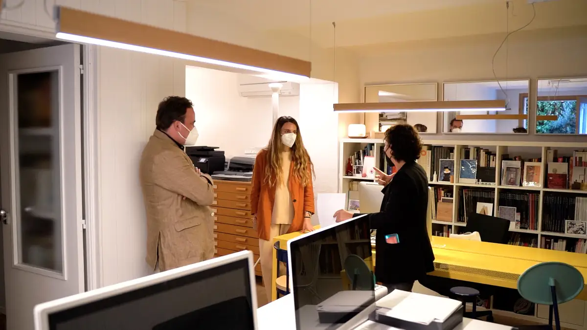 Los dos arquitectos conversan de pie en una oficina con estanterías de libros y una mesa amarilla