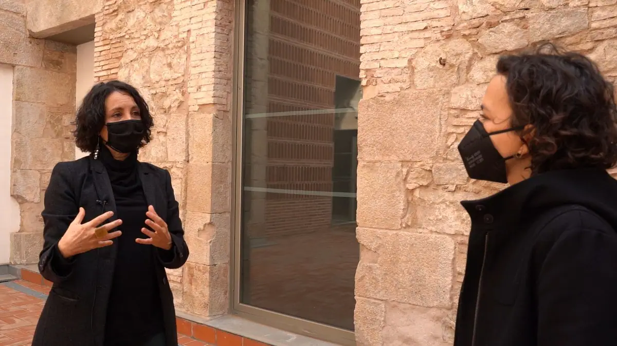 Meritxell Inaraja, con mascarilla, gesticula mientras conversa en un patio con muros de piedra y un gran ventanal