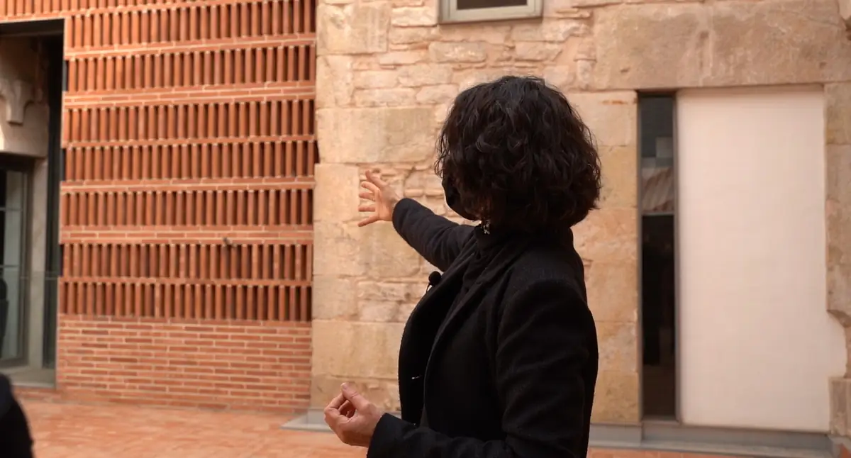 Meritxell Inaraja, de espaldas, señala con la mano una celosía de ladrillo junto a un muro de piedra