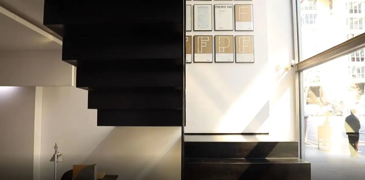 Escalera de peldaños negros volados junto a una pared con carteles enmarcados de los Premios FAD y un gran ventanal a la calle