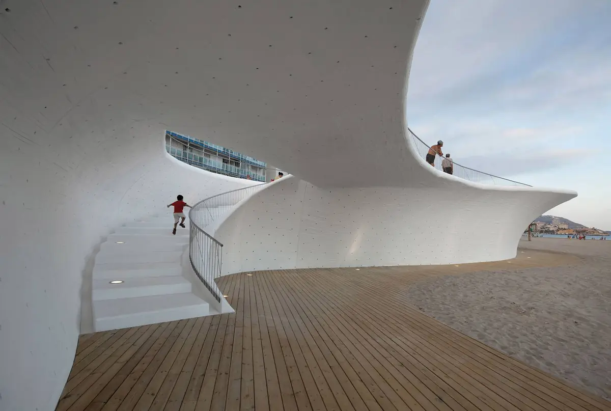 Pasarela de madera junto a la playa bajo una lámina curva de hormigón blanco con una escalera que sube al paseo