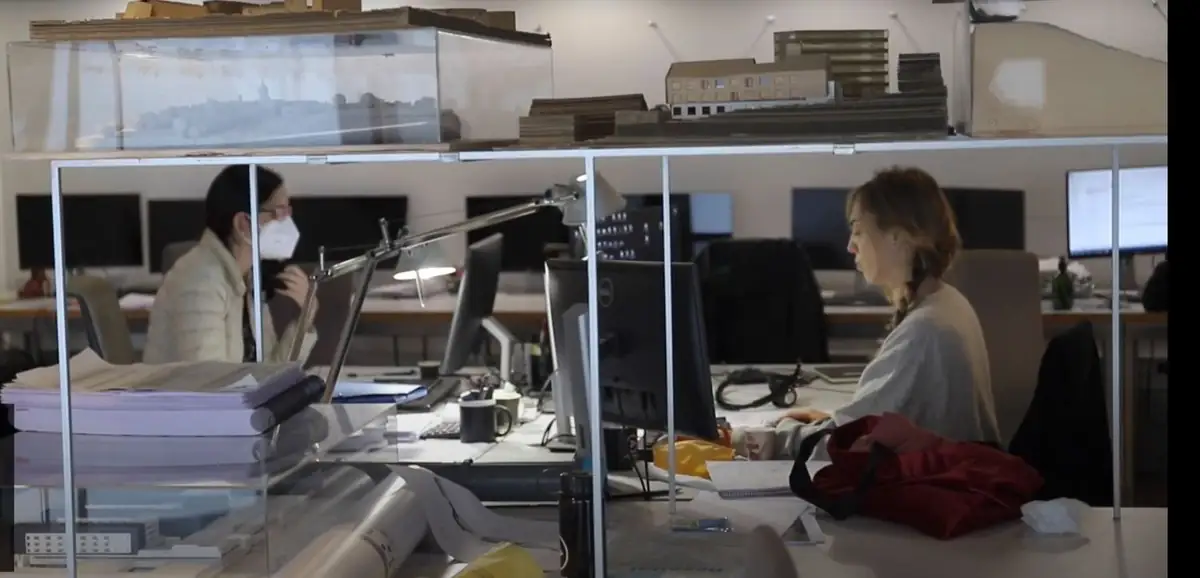 Dos arquitectas trabajando en el estudio tras una estantería de vidrio con maquetas de proyectos