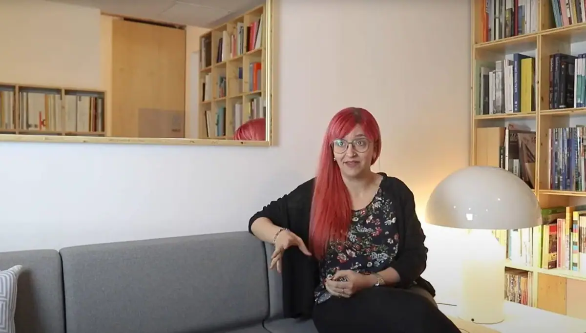 Joana Cornudella, sentada en un sofá gris junto a una estantería y una lámpara blanca, habla durante la entrevista