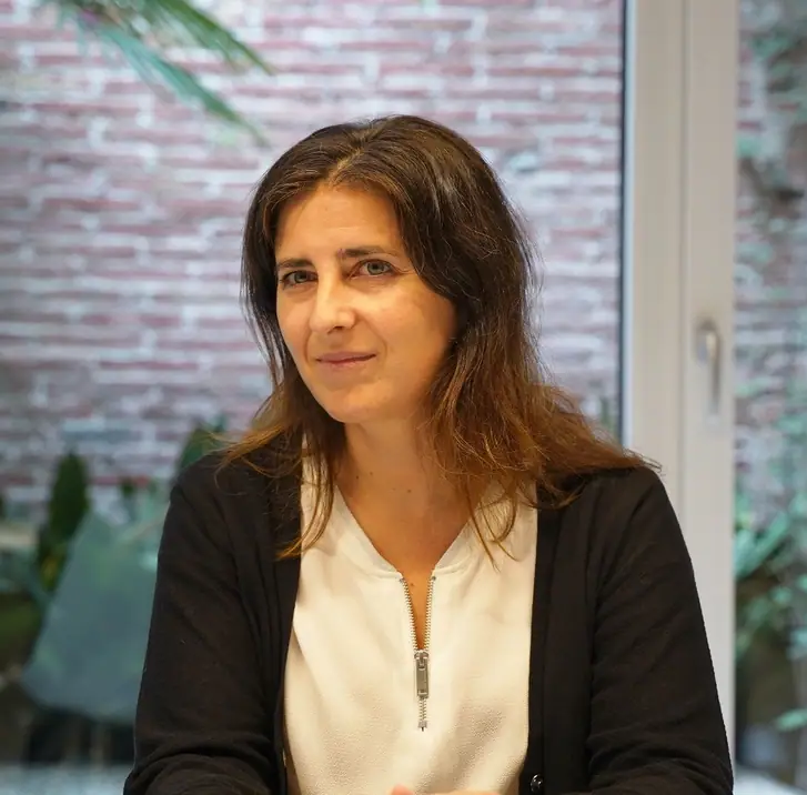 Retrato de Marta Pascual durante la entrevista, con chaqueta negra y blusa blanca, ante una pared de ladrillo