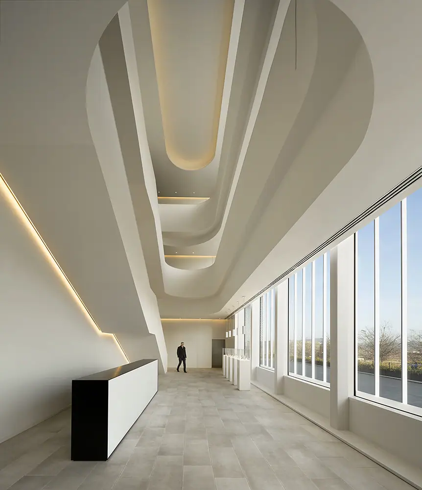 Vestíbulo de techo alto con vacíos curvos iluminados, mostrador blanco y negro y un gran ventanal lateral
