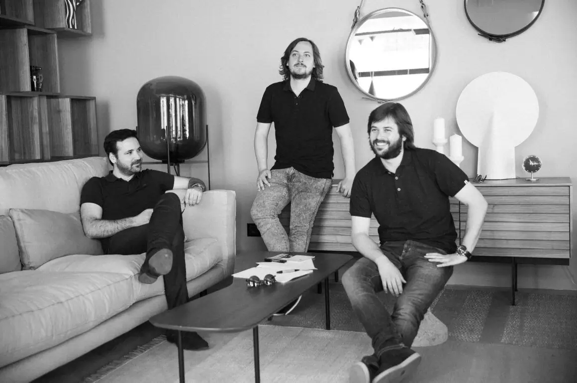 Retrato en blanco y negro de los tres socios de Zooco Estudio en un salón, dos sentados y uno apoyado en un aparador