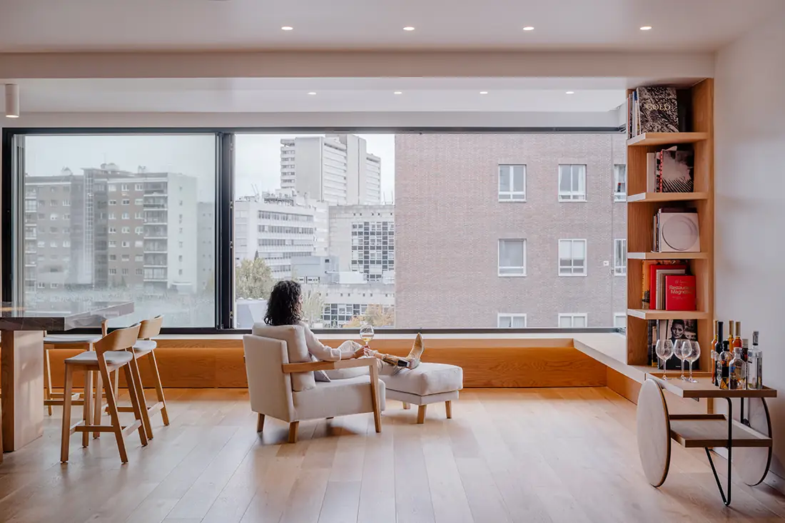 Salón de Casa Paco con suelo de roble, banco corrido bajo un gran ventanal y vistas a los edificios de Madrid