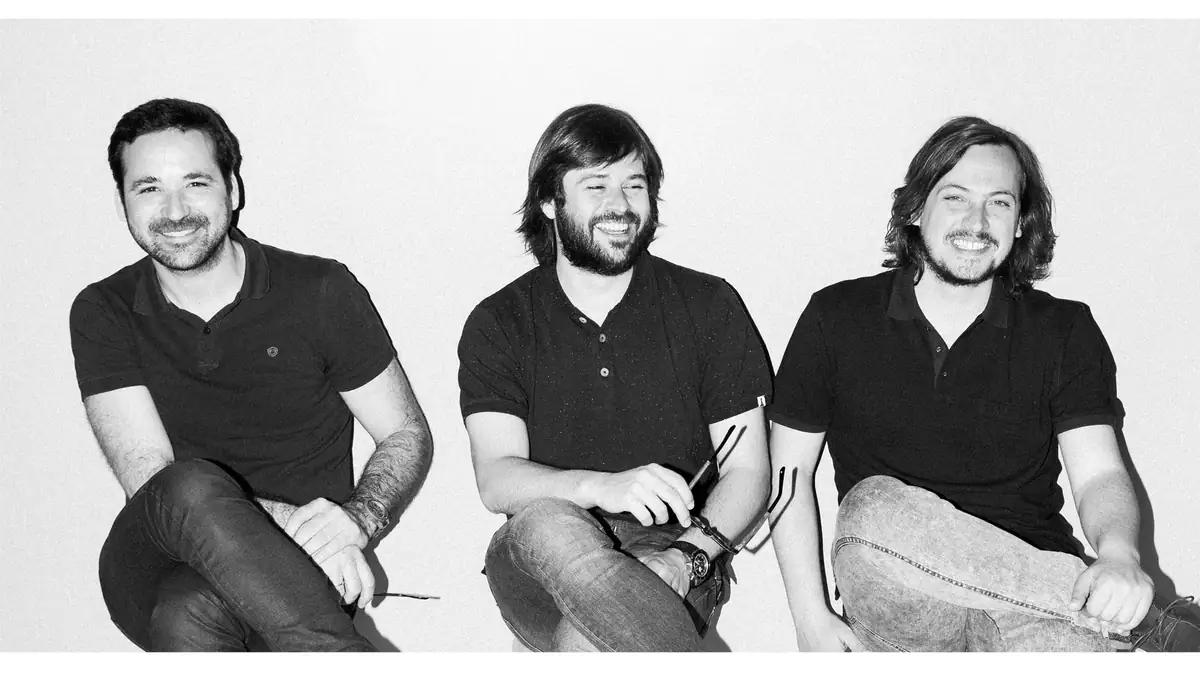 Retrato en blanco y negro de los tres socios de Zooco Estudio, sentados y sonrientes ante una pared blanca